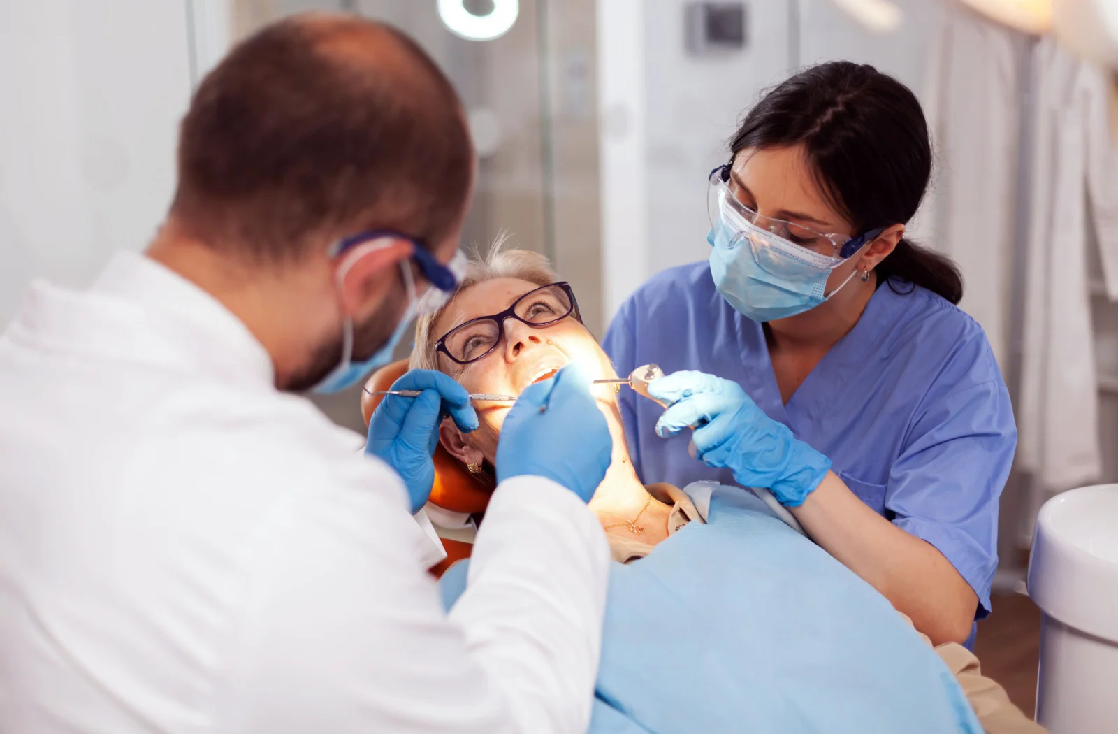 dentist-and-dental-assistant-treating-elderly-patient-in-clinic Dentist and dental assistant treating an elderly patient in a dental clinic, using examination tools and suction device.