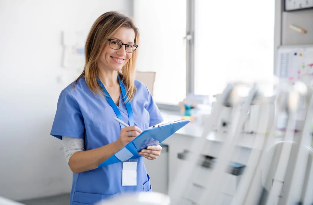 Smiling dental assistant wearing blue scrubs and glasses writing on a clipboard in a dental office.