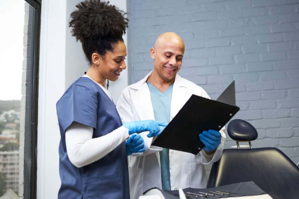 A dentist and a dental assistant wearing scrubs and gloves review a patient chart together while smiling in a dental clinic.