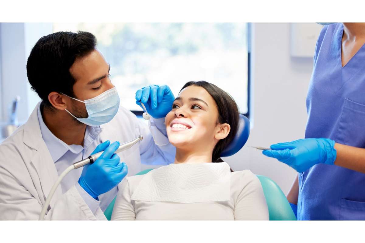 A dentist wearing a mask and gloves examines a smiling patient in a dental chair while a dental assistant stands nearby holding instruments.