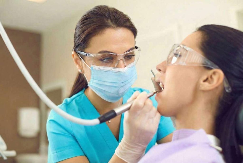 Female dental hygienist in turquoise scrubs, protective eyewear, and blue mask examining a patient's mouth with a dental mirror during a routine dental examination in a modern dental office.