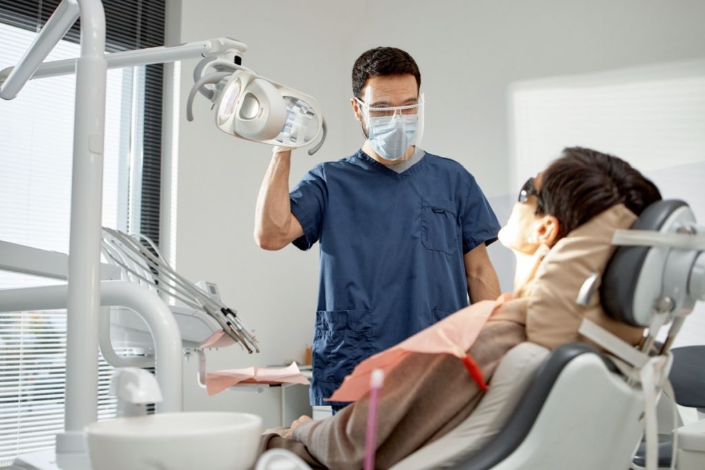 Dentist in navy blue scrubs wearing surgical mask and protective eyewear adjusting overhead dental light while patient reclines in dental chair wearing protective glasses.