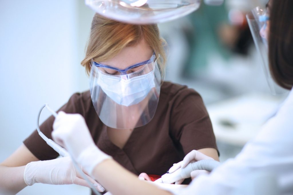 Dental professional wearing protective face shield, mask, and gloves assisting with patient treatment in dental operatory.