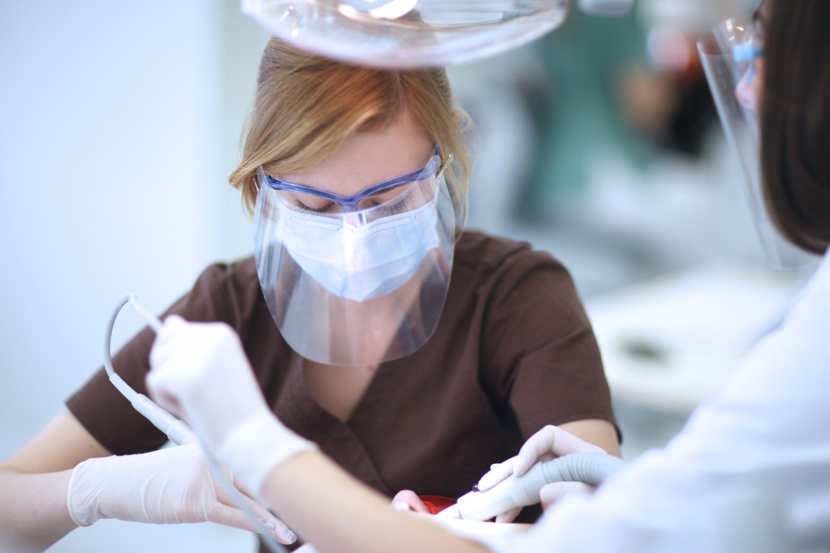 dentist and assistant with mask glasses and splatter screen Dental professional wearing protective face shield, mask, and gloves assisting with patient treatment in dental operatory.