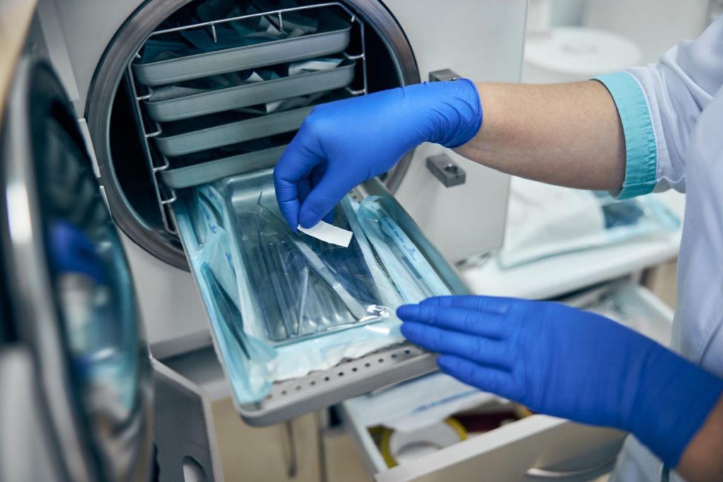 Gloved hands placing sealed dental instruments into an open autoclave sterilization machine in a clinical setting.